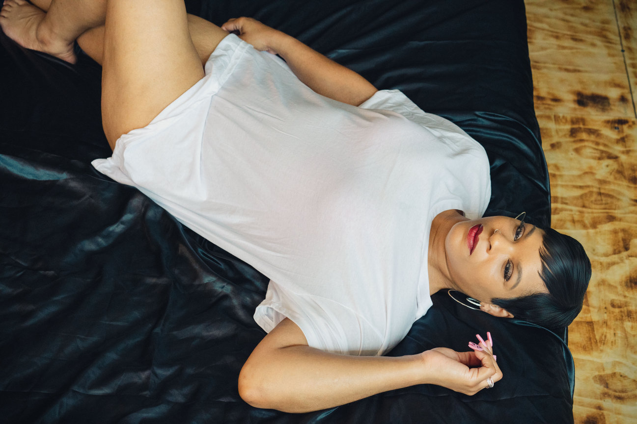 A woman in a white t-shirt reclines on a black bedspread, looking directly at the camera.