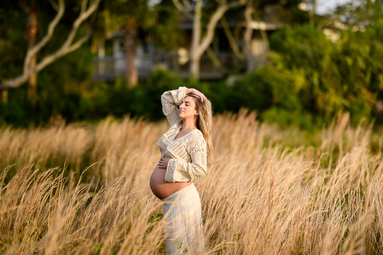Pregnant woman in a field of tall grass, wearing a light-colored dress, surrounded by greenery.