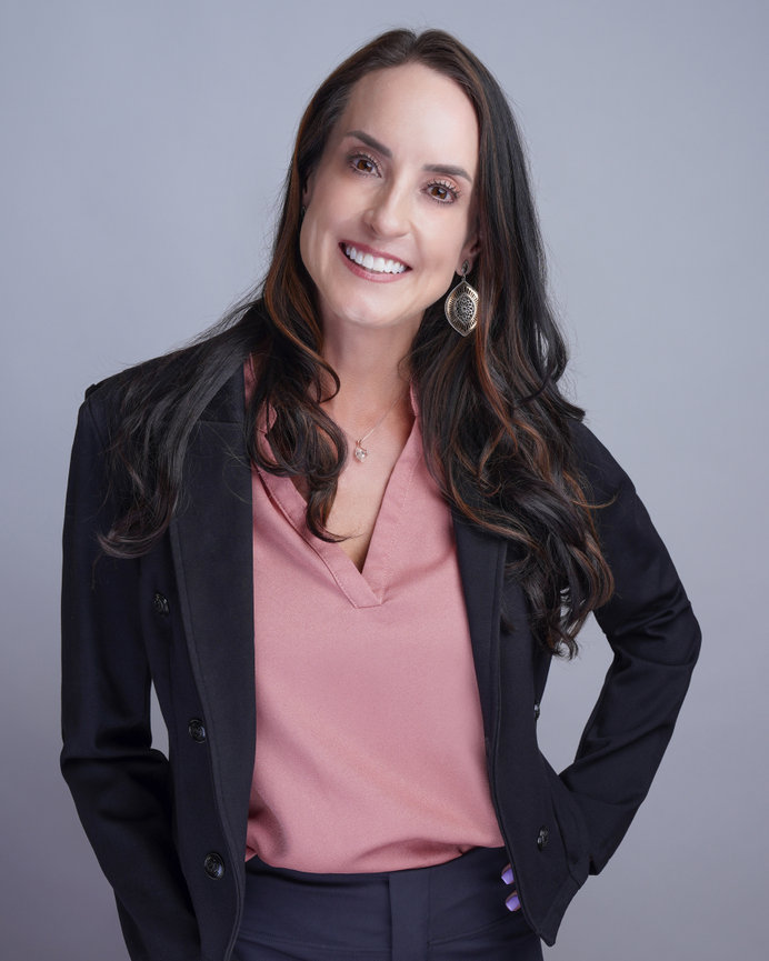 Woman smiling, wearing a black blazer and pink blouse, with long hair and hoop earrings, standing against a gray background.