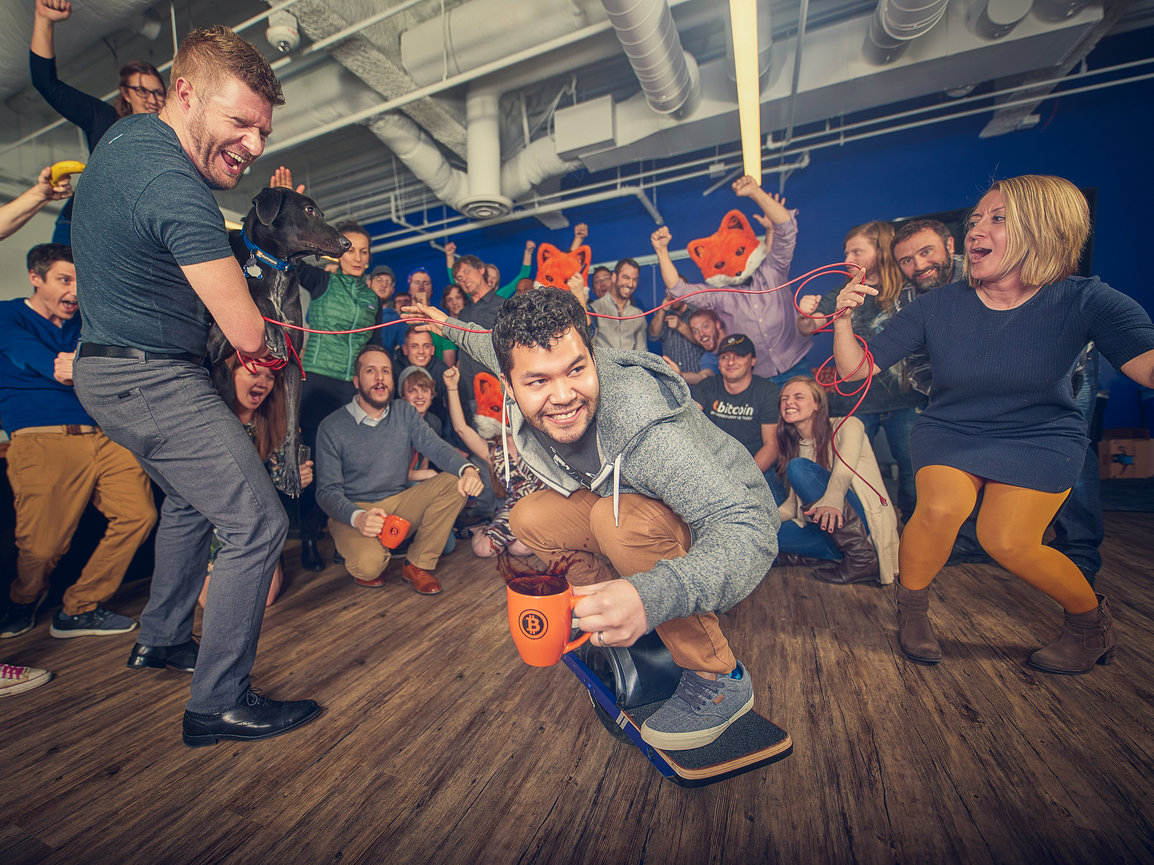 A group of people in an office setting celebrating humorously, with one person balancing on a board holding an orange mug.