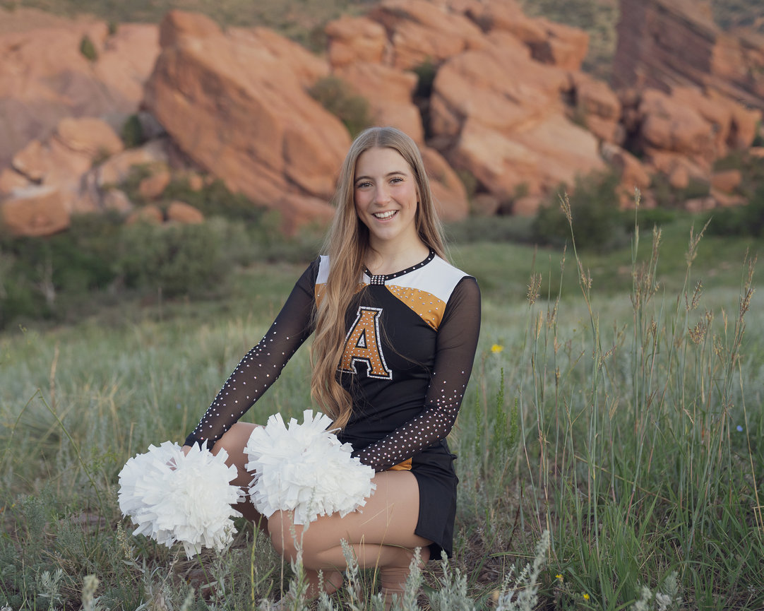 A cheerleader in uniform holding pom-poms, kneeling in a grassy field with large rock formations in the background.