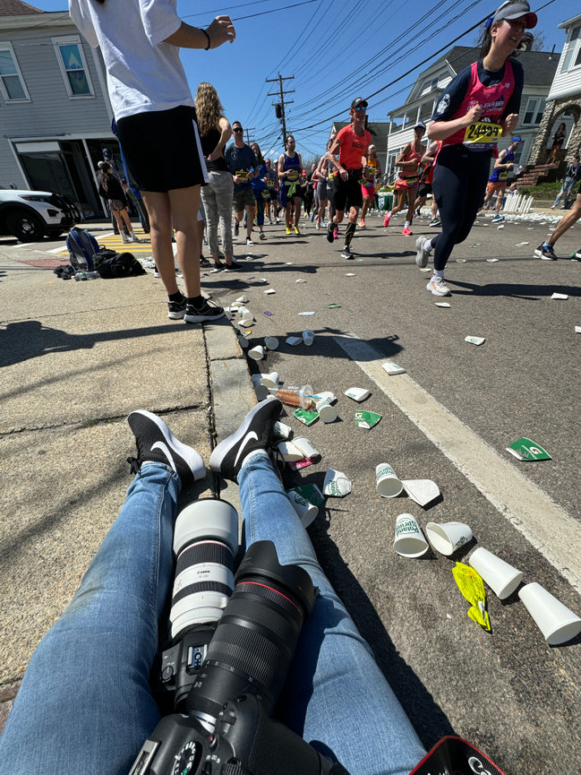 Paula Swift sitting on sidewalk with cameras, photographing the Boston marathon. Runners and discarded cups on the street.