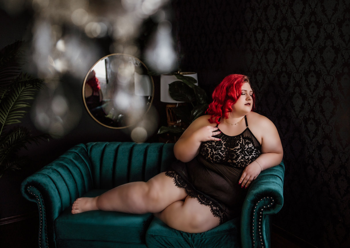 Curvy boudoir Port Angeles, WA Woman with red hair in lace dress lounging on a teal sofa, with a round mirror and dark wallpaper in the background.