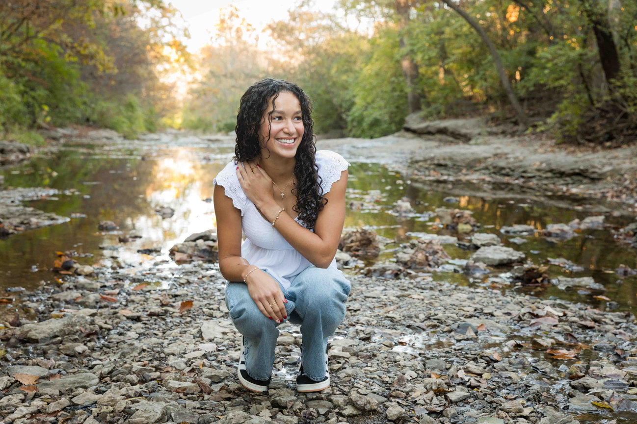 Woman crouching on a rocky riverbed in a forest, smiling, wearing a white top and jeans, surrounded by trees.