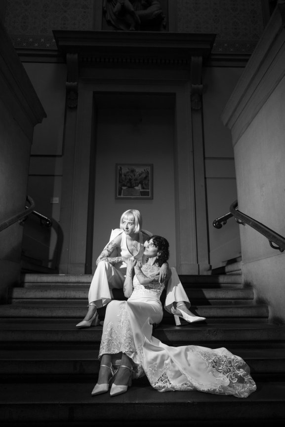 High-contrast black and white editorial wedding portrait of two brides on a staircase at Manchester Art gallery, one in a tailored trouser suit and the other in a long lace dress, captured with direct flash.