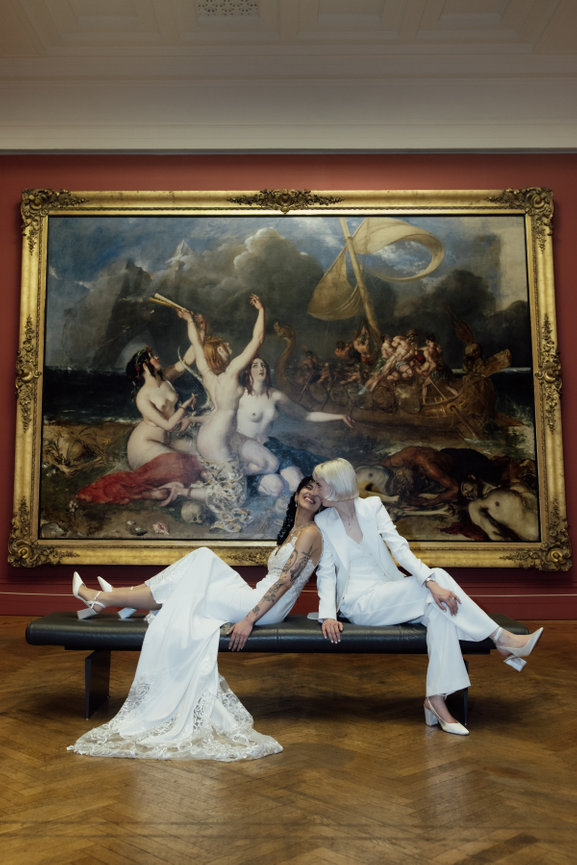 Editorial wedding portrait of two brides—one in a white trouser suit and the other in a lace dress—sitting on a bench before a grand classical painting at Manchester Art Gallery.