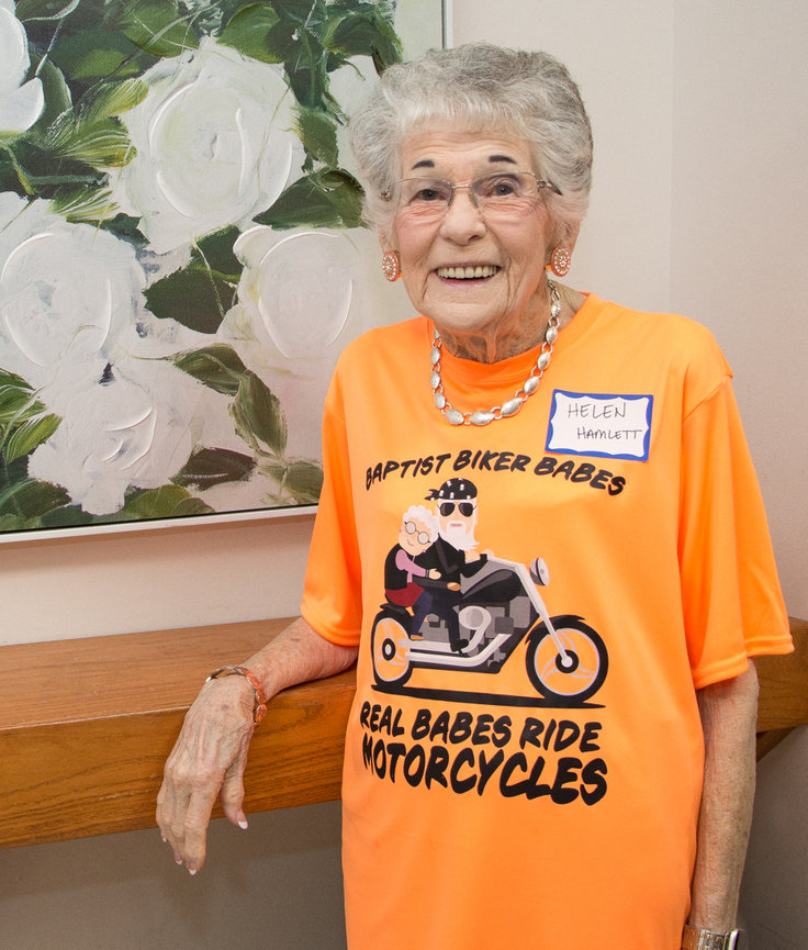 Elderly woman smiling, wearing an orange biker-themed T-shirt, standing next to a painting of white flowers.