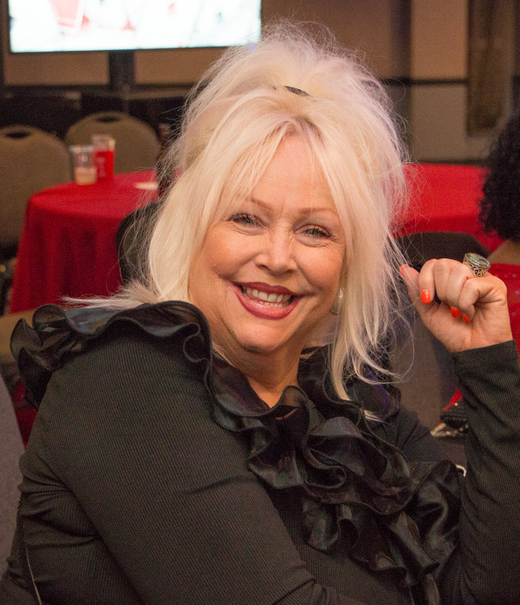 Smiling woman with blonde hair and black ruffled top, seated at a table with a red tablecloth in a social setting.
