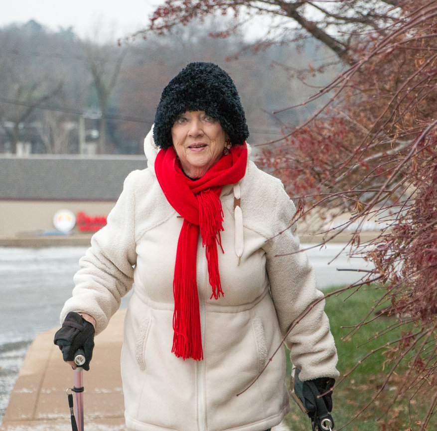 Elderly woman in a winter coat and red scarf, using a walker on a snowy path.