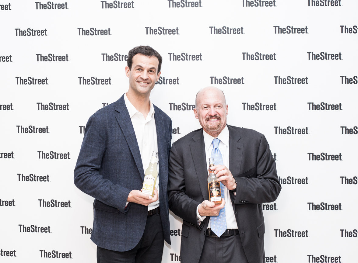 Two people holding bottles, smiling, in front of a backdrop with TheStreet logo.