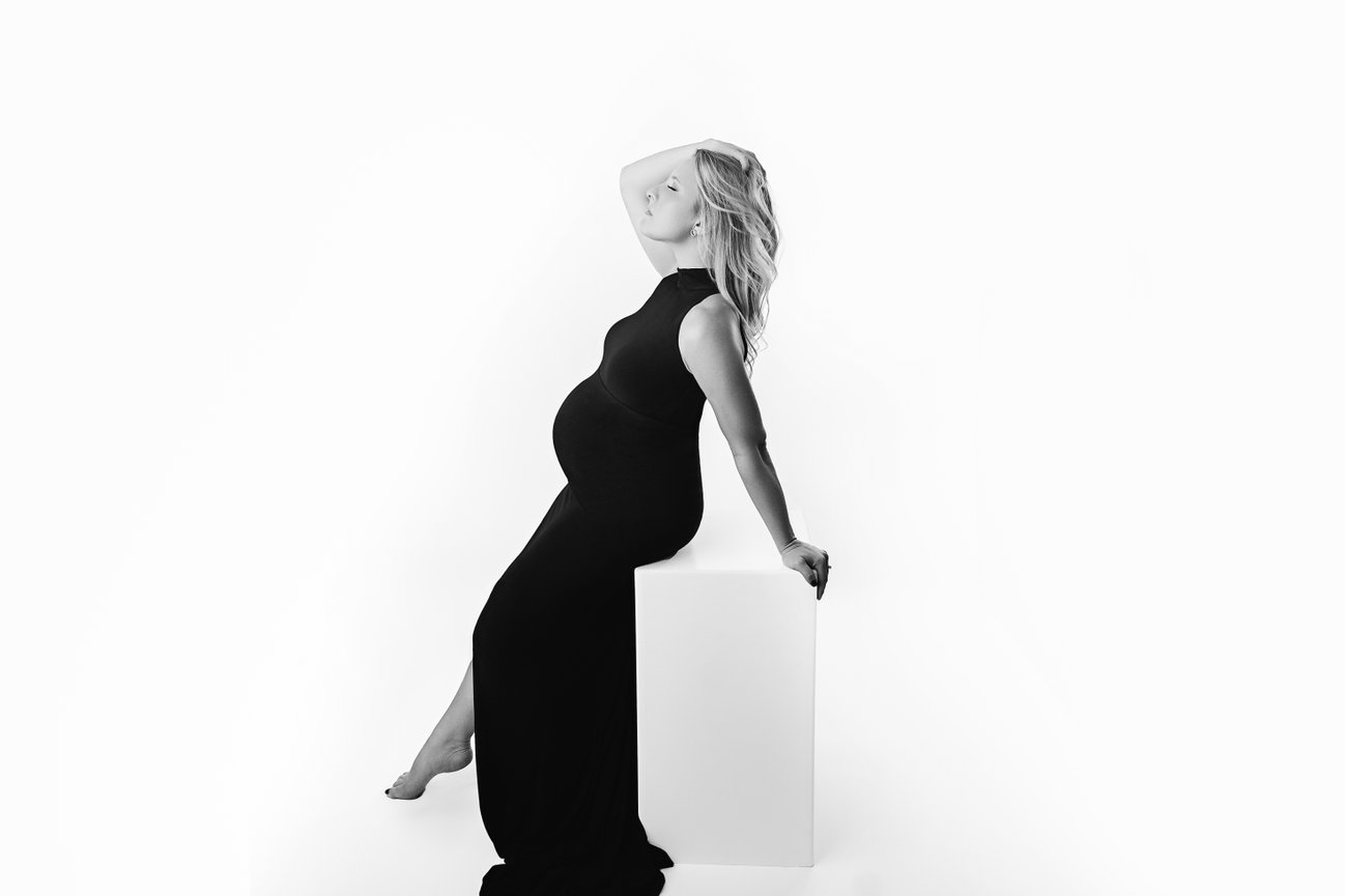Black and white maternity image in white studio, featuring a fitted black gown with slit and refined posing.