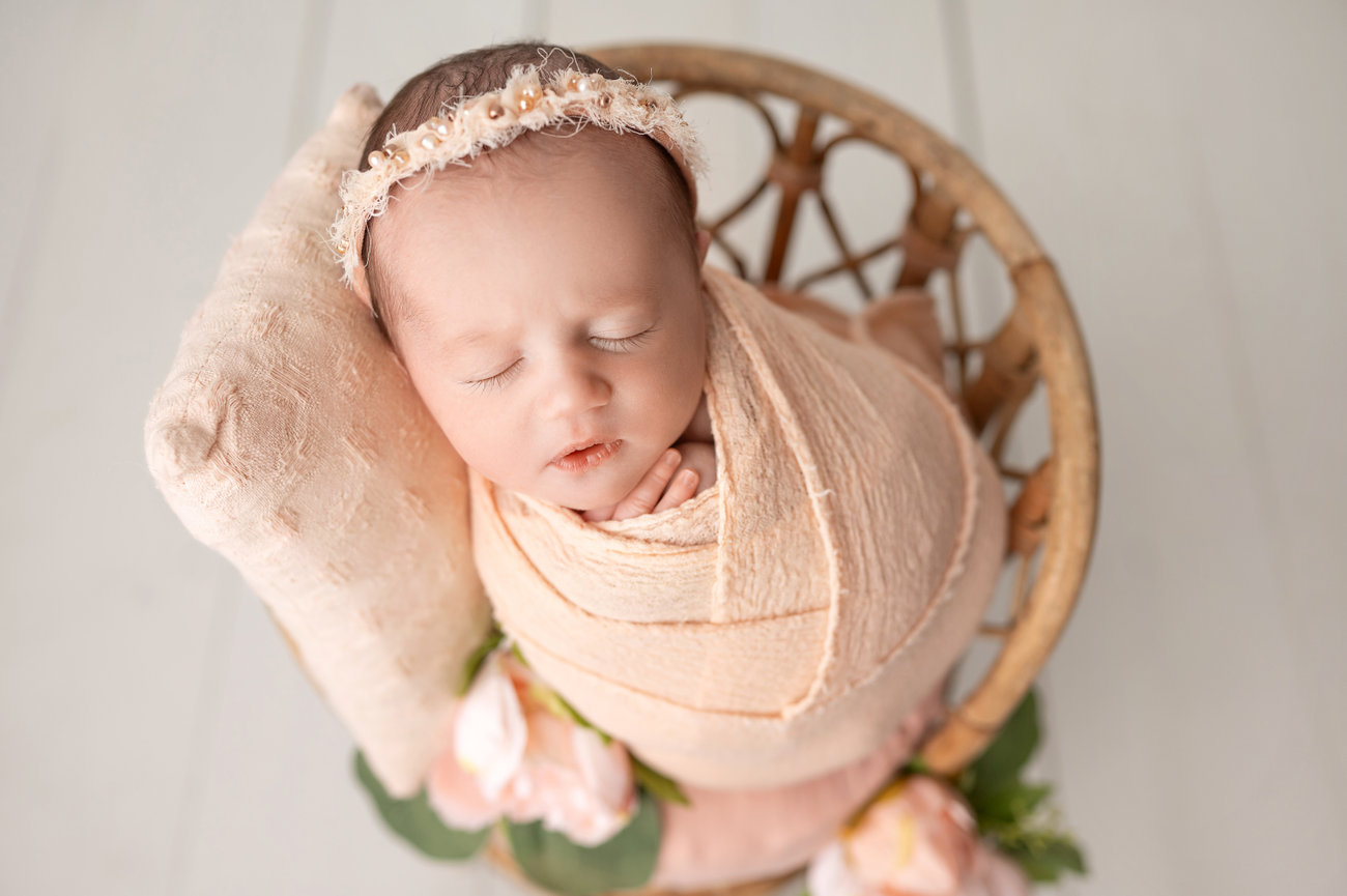 Newborn baby swaddled in textured cheesecloth wrap, sleeping peacefully in a wooden chair with flowers during studio session in Manasquan, NJ