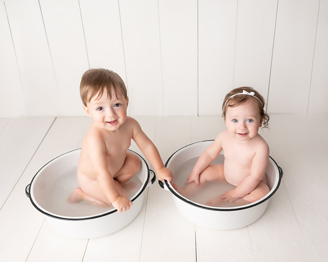 Twin one year old babies laughing while rinsing off frosting in white bathtubs