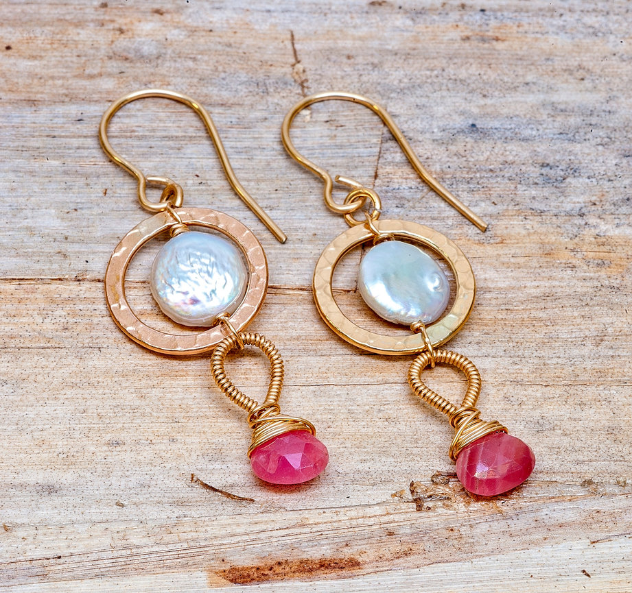 Pair of gold dangle earrings featuring hammered circular frames with iridescent coin pearls in the center, and wrapped wire loops holding faceted pink teardrop gemstones, displayed on a rustic wooden surface.