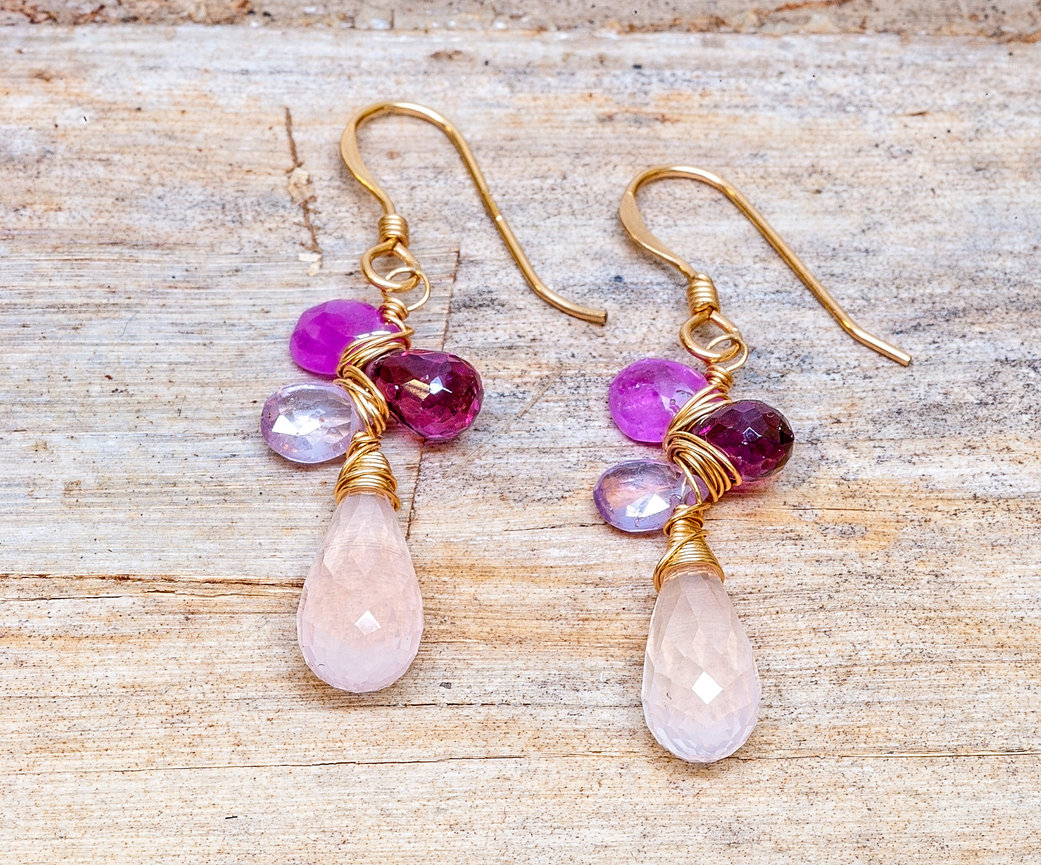Pair of gold-wire hook earrings with faceted pale pink teardrop gemstones topped with clusters of pink, purple, and deep magenta beads, displayed on a rustic wooden surface.