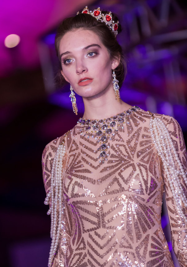 Fashion model on the runway wearing a sparkling, geometric-patterned gown adorned with pearl strands on the sleeves, a jeweled necklace, long dangling earrings, and a crown with red gemstones, set against a purple-lit background.