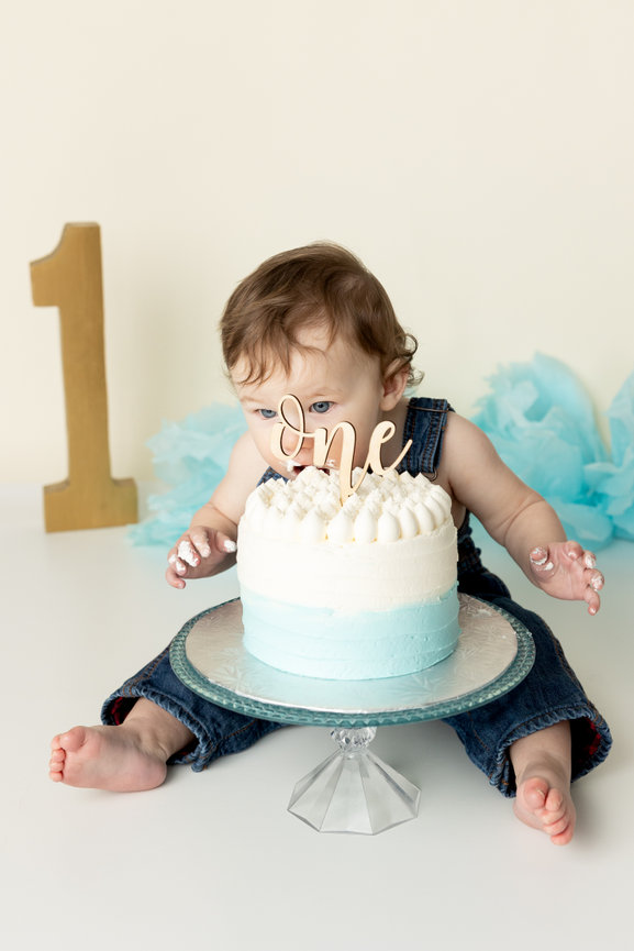 Baby in overalls with a one cake on a glass stand, large number one prop in the background.