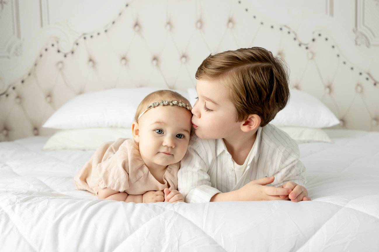 Two young children on a bed; an older child kissing a baby on the head.