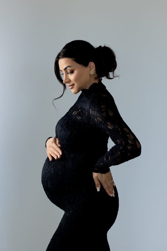 Pregnant woman in a black lace dress, smiling and gently holding her belly against a neutral background.