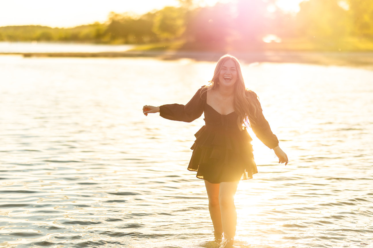 Woman in a black dress laughing and standing in sunlit water at sunset.