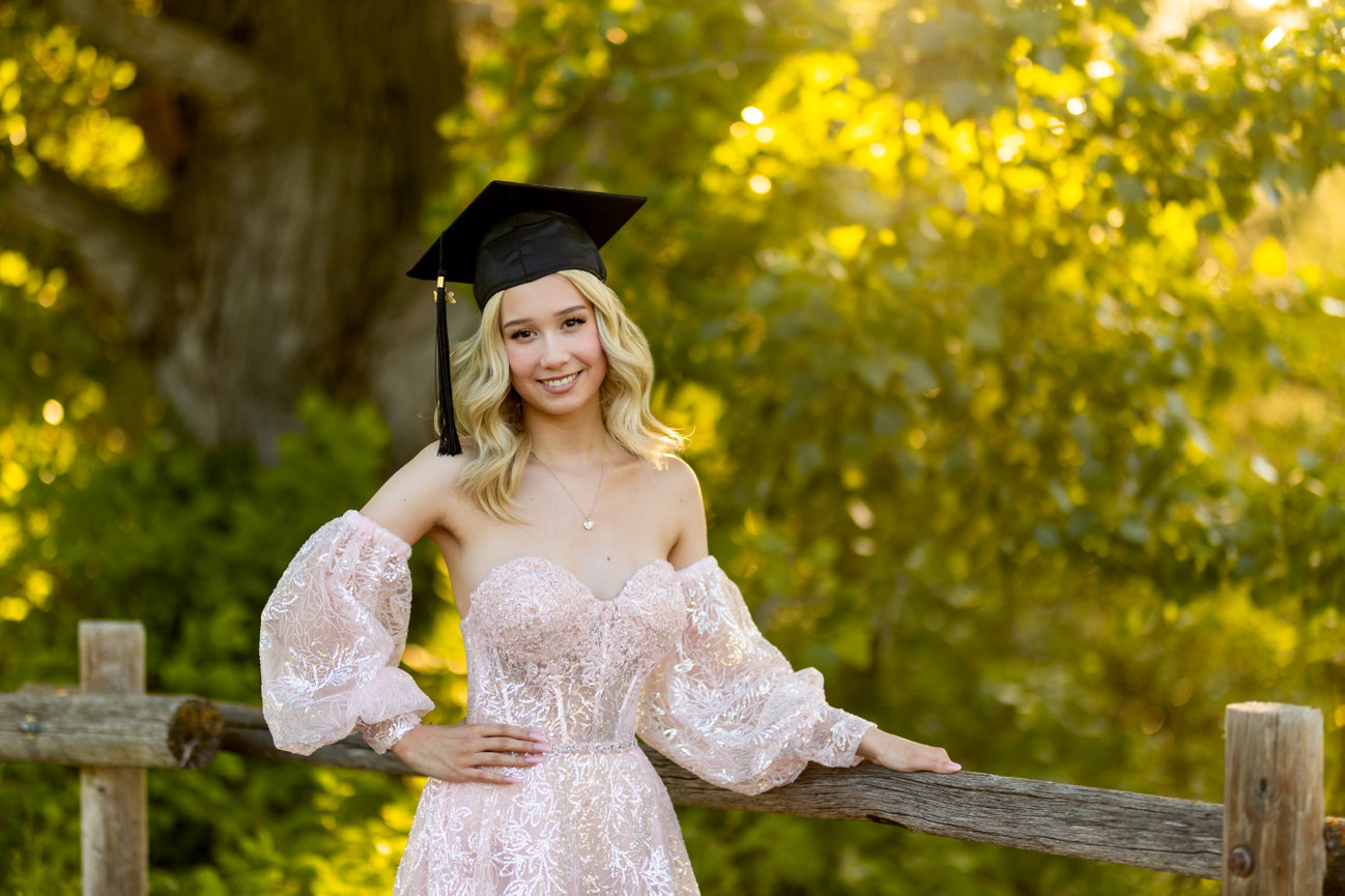 Graduate in a pink dress and cap smiling, leaning on a wooden fence with lush greenery in the background.