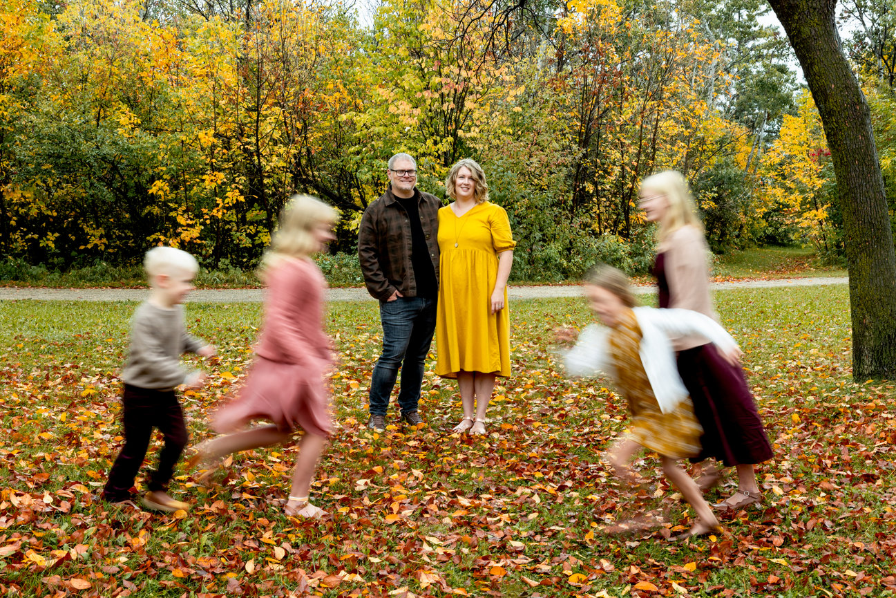 A couple stands smiling in a park with autumn foliage as children in motion blur play around them.