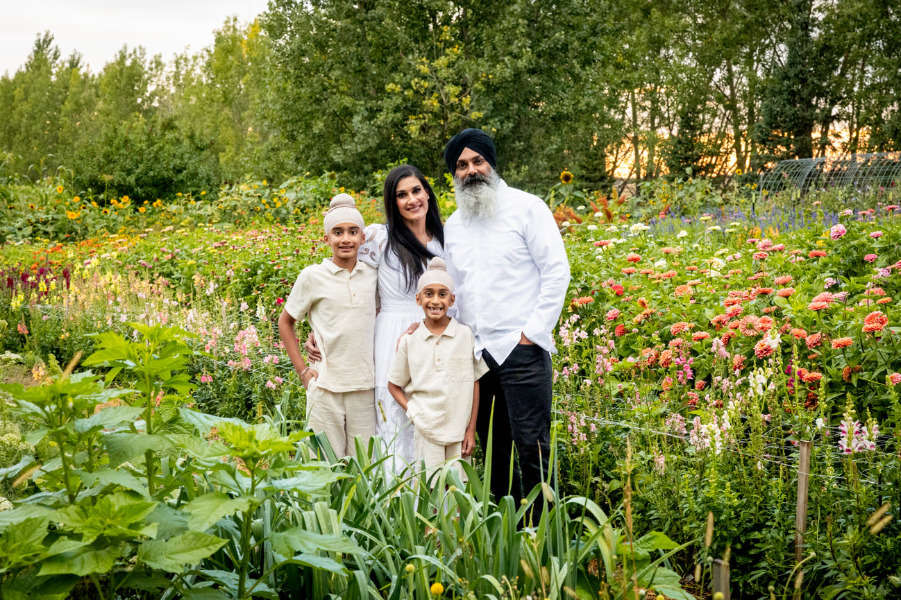 A family of four stands in a lush garden surrounded by various plants and flowers, smiling at the camera.