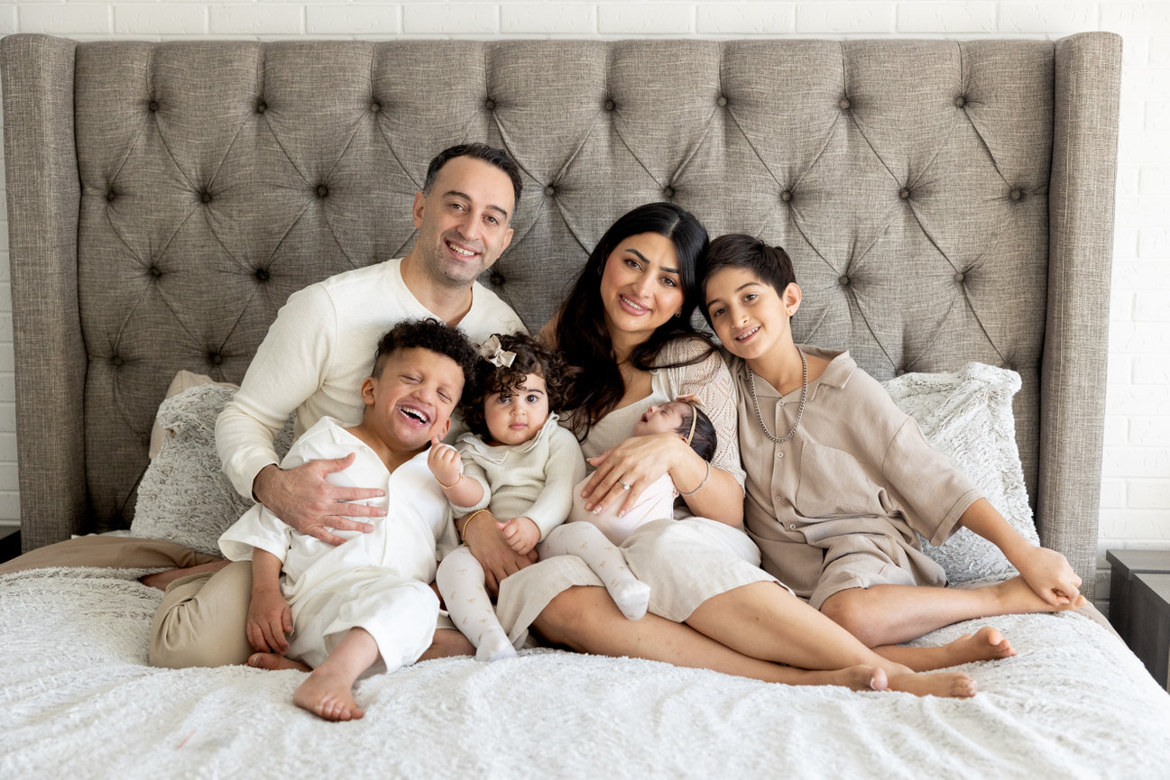 A family of five sitting on a bed, smiling. Parents with three young children, in a cozy, neutral-toned setting.