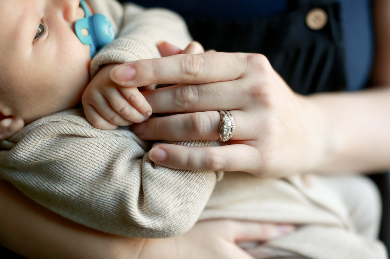 Adult holding baby in tan outfit, baby has a blue pacifier. The adult's hand has a sparkly ring.