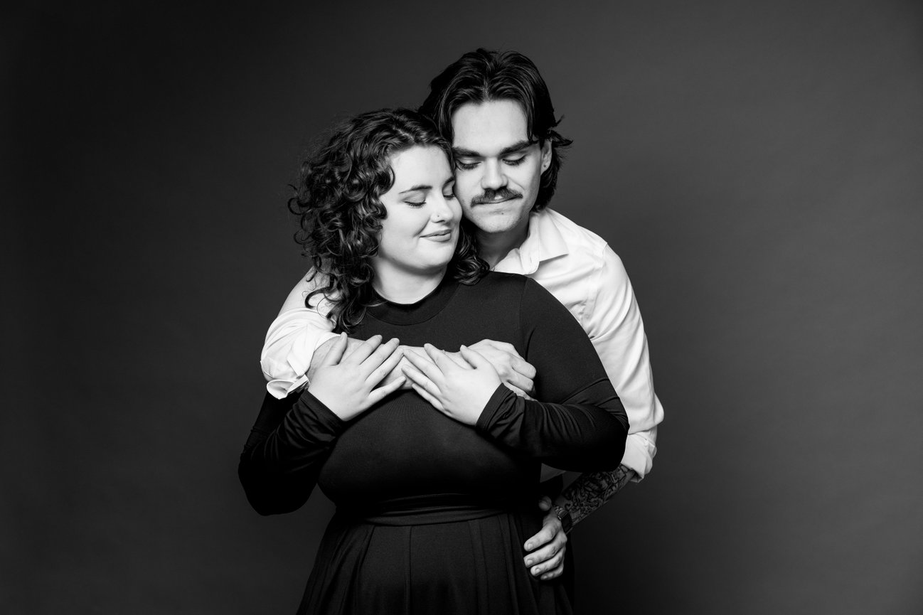 Man embracing woman from behind in a black-and-white portrait with a neutral background.