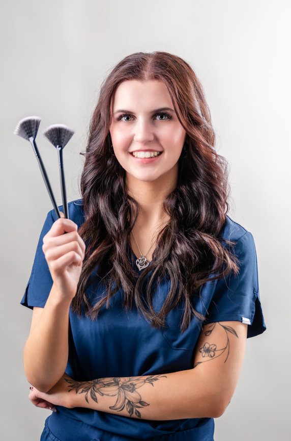 Woman in blue scrubs holding two make-up brushes looking at the camera smiling