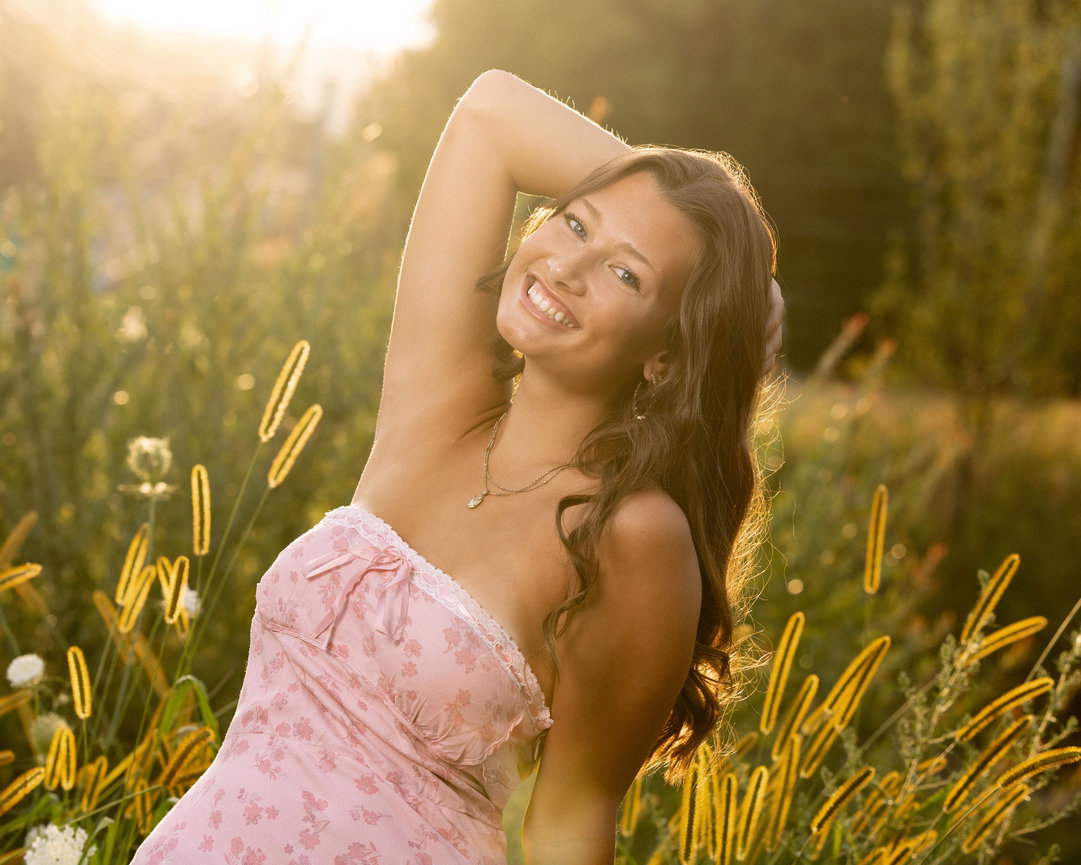 High School senior portrait of blonde girl smiling with hand in hair as the sun sets behind her in a field