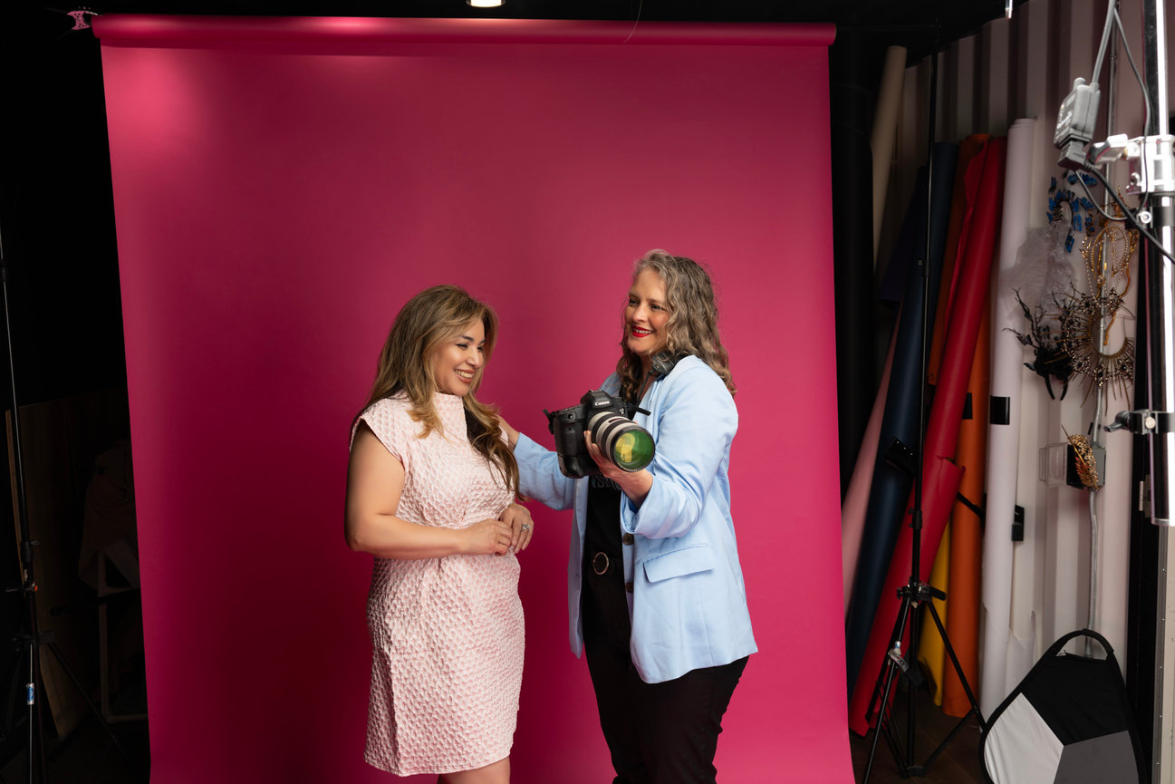 Two women pose in front of a pink backdrop; one holds a camera, while the other smiles.