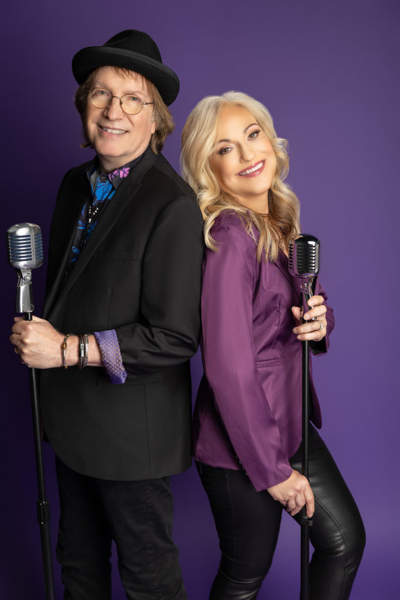 Branding headshot of two professionals posing with microphones in coordinated purple outfits against a purple background.