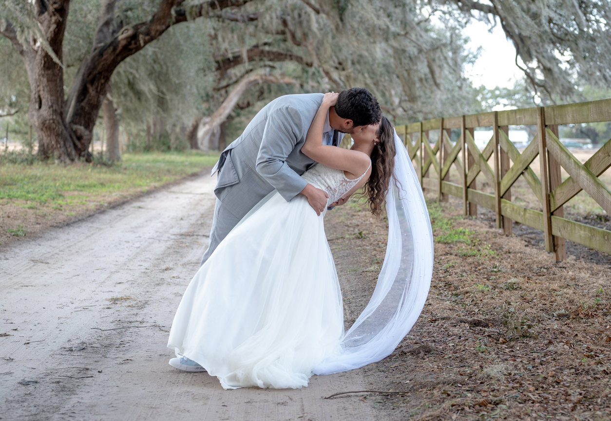 Bride and groom kissing beside a rustic path with a long veil trailing behind.