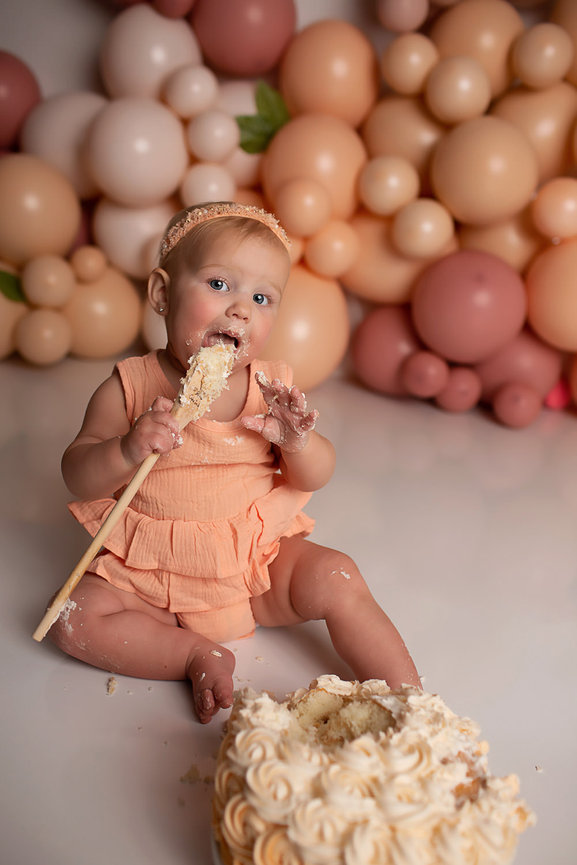 Baby girl eating cake off a wooden spoon peach pink and neutral colored balloons in the background during her first birthday cake smash photo session with Kansas City photographer Dreamworks Photography 