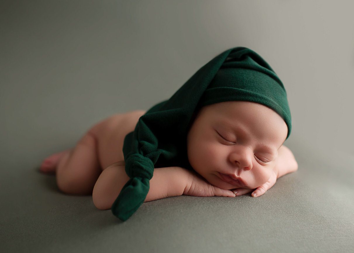 Newborn baby sleeping on his belly on sage green fabric with dark green sleepy cap with his hands under his chin and feet behind him from photo session with newborn photographer Dreamworks Photography in Kansas City 