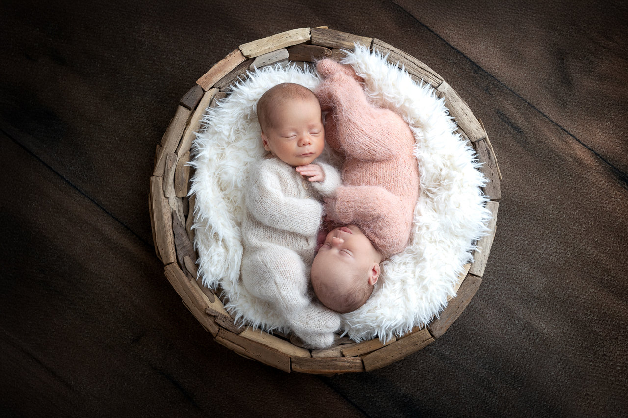 Two newborn babies are peacefully sleeping in a cozy driftwood basket on a wooden floor during their newborn photo session with Catherine Whitney Photography in St. Augustine, Florida