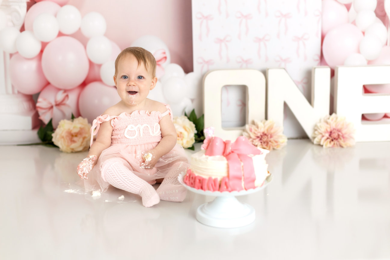 A baby girl in a pink dress sits before a beautifully decorated cake, her expression full of delight during her cake smash photo session with Catherine Whitney Photography