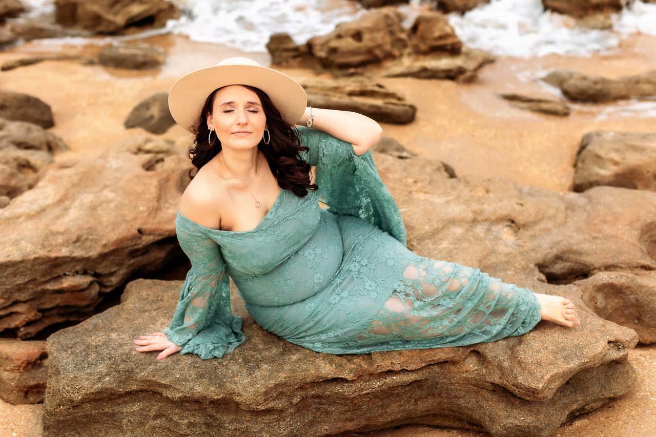 Woman in a flowing teal dress and hat reclining on rocks by the water during a maternity photography session with Catherine Whitney Photography in St. Augustine Florida