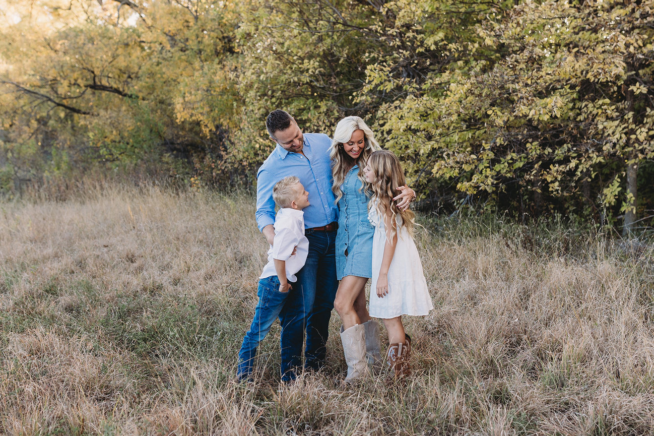 Family of four in a grassy field with trees in the background, smiling and wearing casual blue and white outfits.