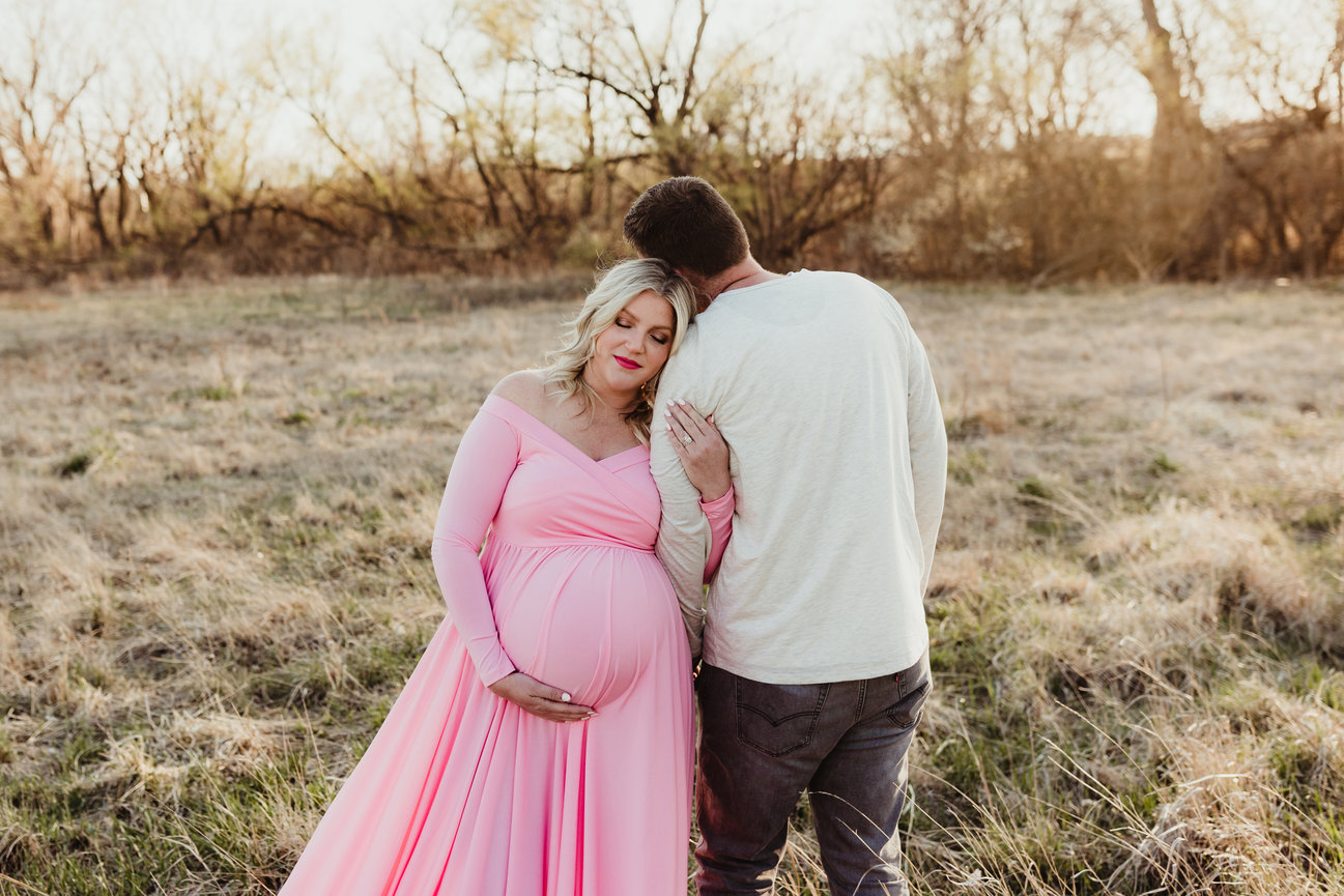 Pregnant woman in a pink dress leans on a man's shoulder in a grassy field.