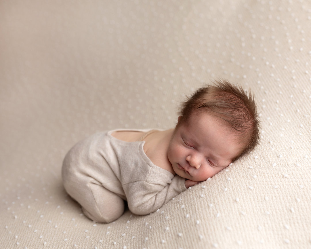 Omaha newborn Photographer photographs sleeping newborn in beige outfit lies on a textured, light-colored blanket.