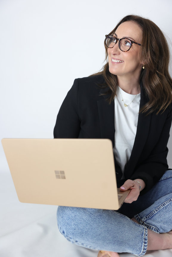 Smiling woman in glasses using a laptop while sitting cross-legged against a white background. melbourne website photographer
