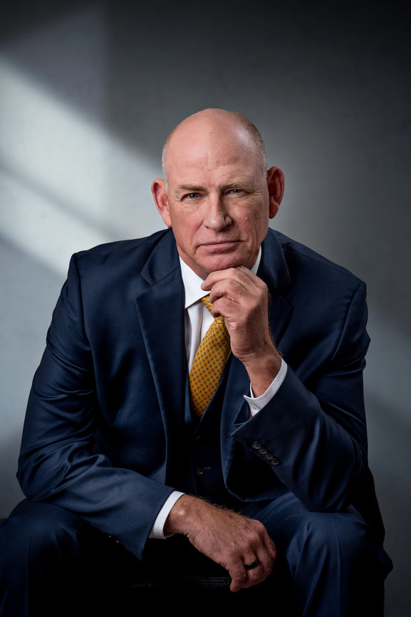 An older man in a suit leaning on his knee with his hand under his chin.