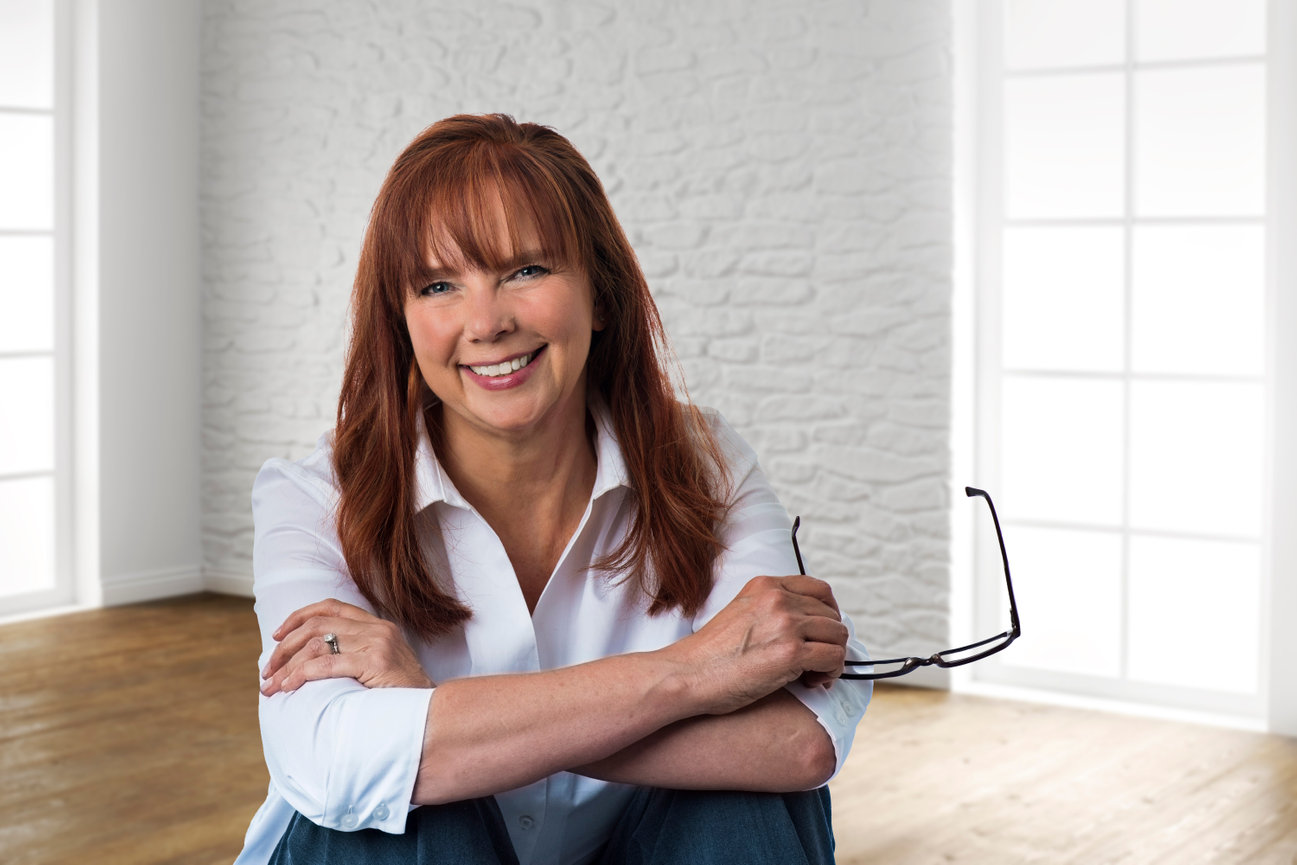 A woman wearing a white shirt sitting with her knees up and arms crossed smiling at the camera.