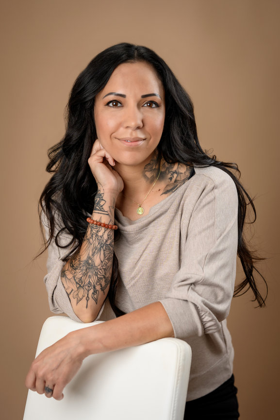 Woman with dark long hair leaning on a chair with her hand by her neck smiling.