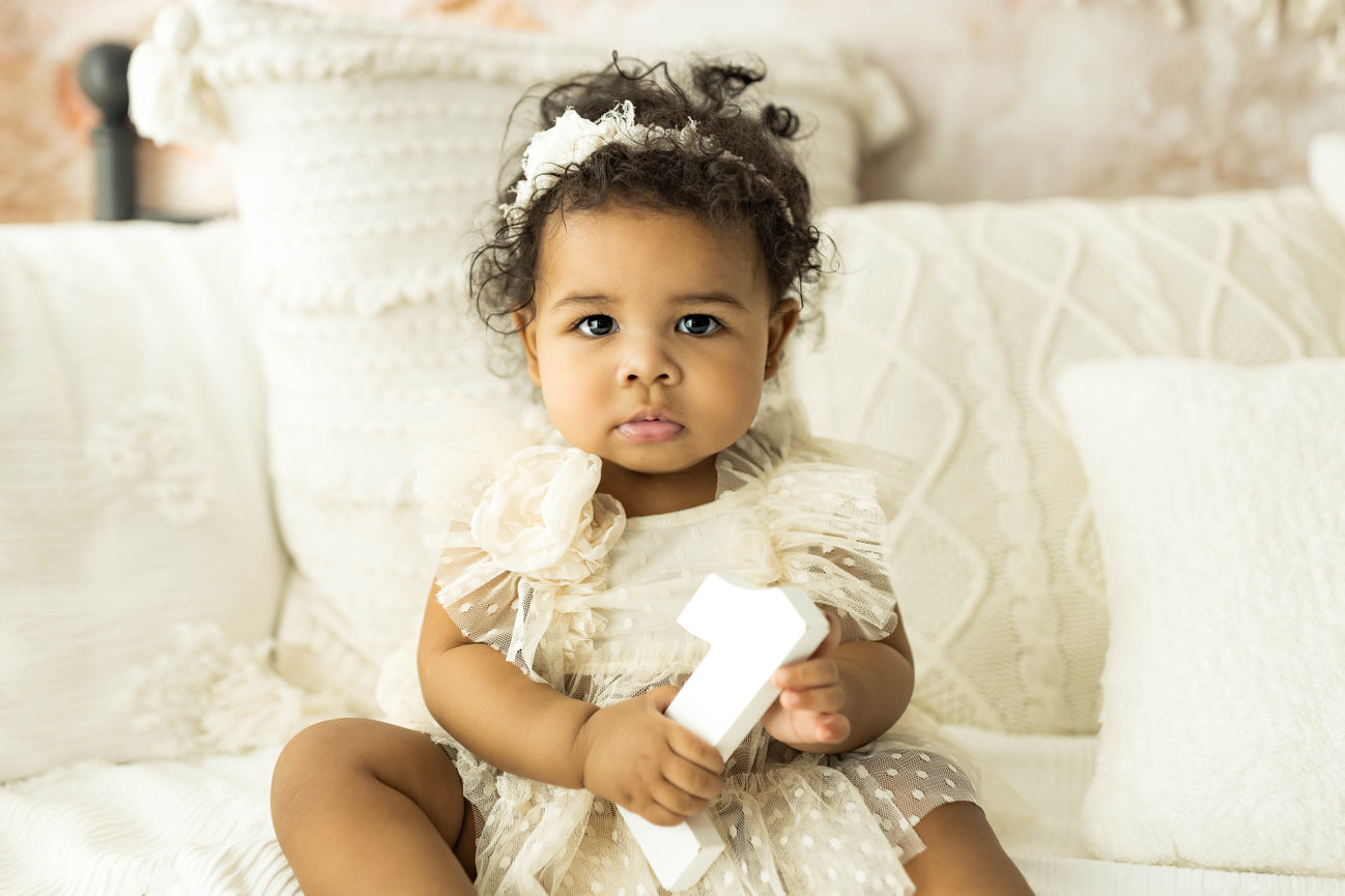 Baby sitting in cream dress against neutral indoor session background during 12-month milestone portrait holding number one