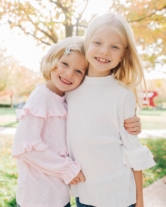 Two young girls hugging and smiling in a sunlit park with autumn trees in the background.