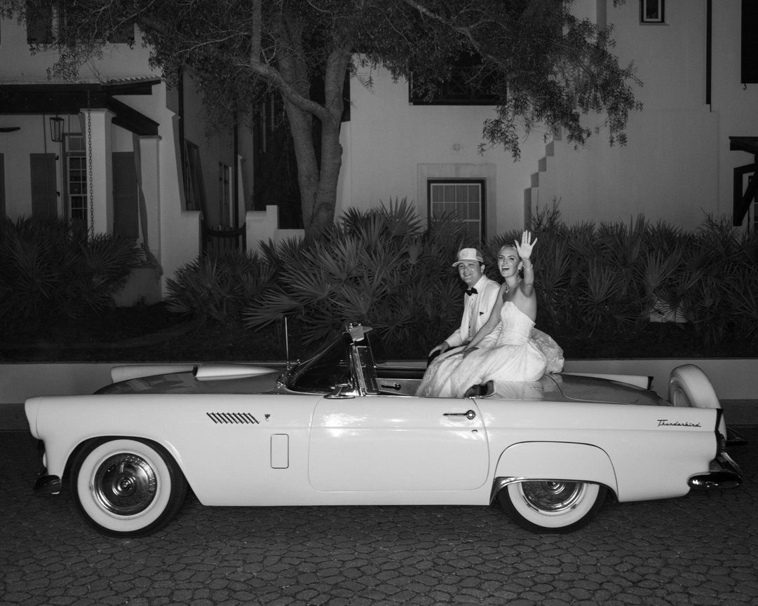 Black and white image of bride and groom sitting on the back of a Thunderbird Convertible in Alys Beach, Florida waving goodbye to guests. Alys Beach wedding photography by Heather Durham.